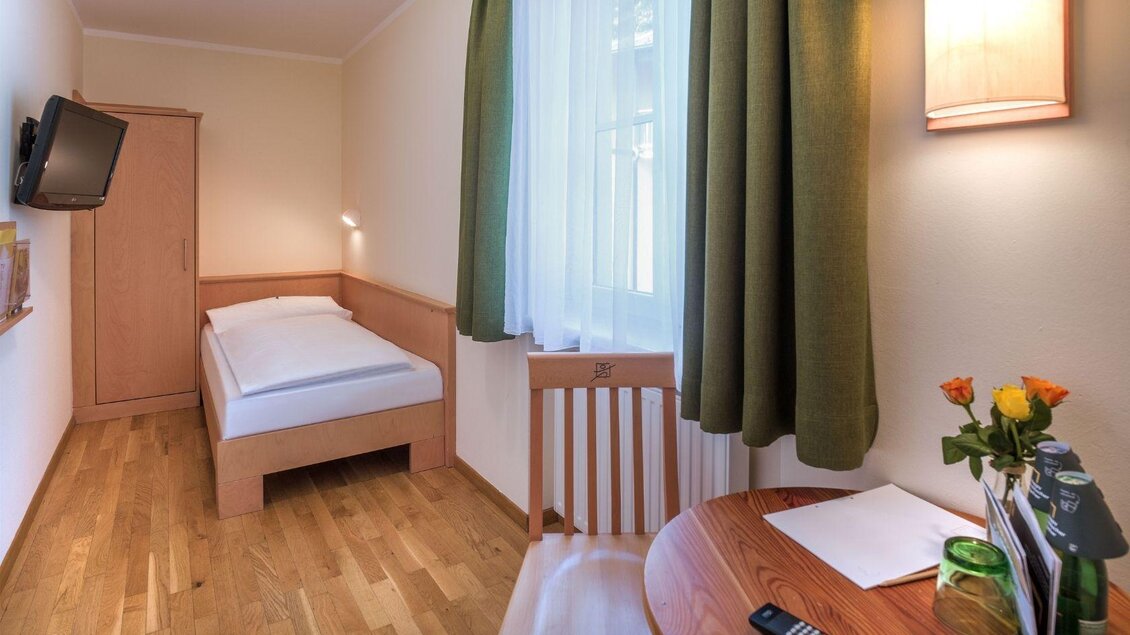 Ein einfaches Zimmer mit einem Einzelbett und einem Schreibtisch. Es gibt einen Fernseher und große Fenster mit grünen Gardinen. | © JUFA Hotels
