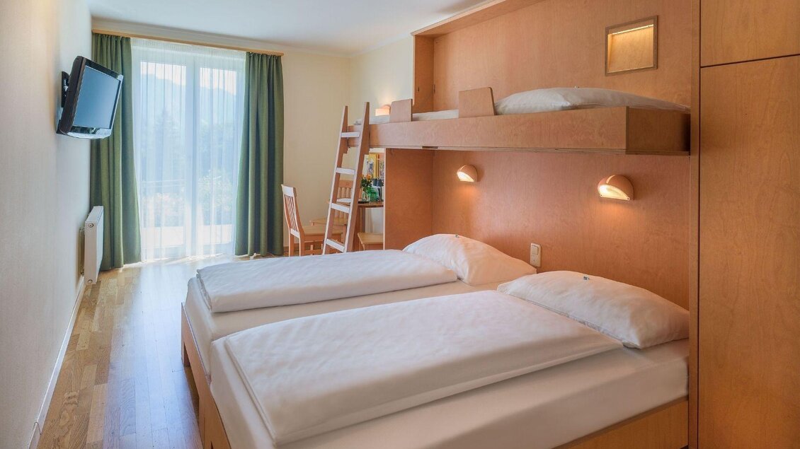 Ein gemütliches Hotelzimmer mit zwei Betten und einem Hochbett. Der Raum hat große Fenster, die viel Tageslicht hereinlassen. | © JUFA Hotels