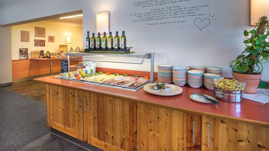 Ein gemütlicher Buffetbereich mit einer Auswahl an frischen Lebensmitteln und Getränken. Auf der Theke sind Obst, Aufschnitt und verschiedene Servierplatten angeordnet. | © JUFA Hotels