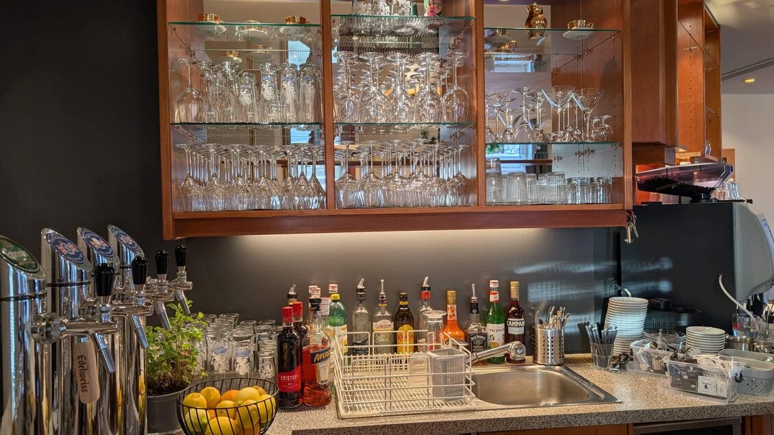 Eine moderne Küche mit einem gut ausgestatteten Barbereich. Gläser und verschiedene Spirituosen sind ordentlich in der Vitrine und auf der Arbeitsfläche angeordnet. | © Sandy`s Café Bar