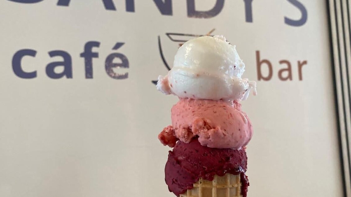 Ein drei­schichtiger Eisbecher in einer Waffel, gehalten von einer Hand. Im Hintergrund ist das Logo von "SANDY'S café bar" zu sehen. | © Sandy`s Café Bar
