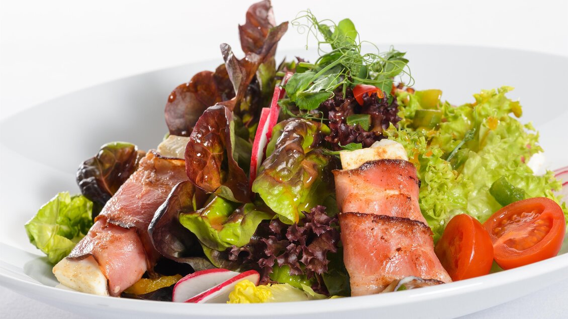 Ein frisch zubereiteter Salat mit verschiedenen Blattsalaten, Kirschtomaten und Radieschen. Dazu gibt es mit Speck umwickelte Zutaten, die das Gericht abrunden. | © Tauroa GmbH