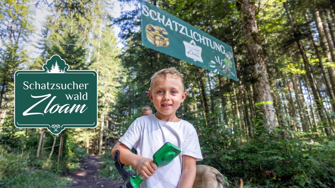 Ein fröhliches Kind steht auf einem Waldweg und hält ein Schatzsuchergerät in der Hand. Im Hintergrund sieht man ein Schild mit dem Aufschrift „Schatzlichtung“. | © www.zloam.at