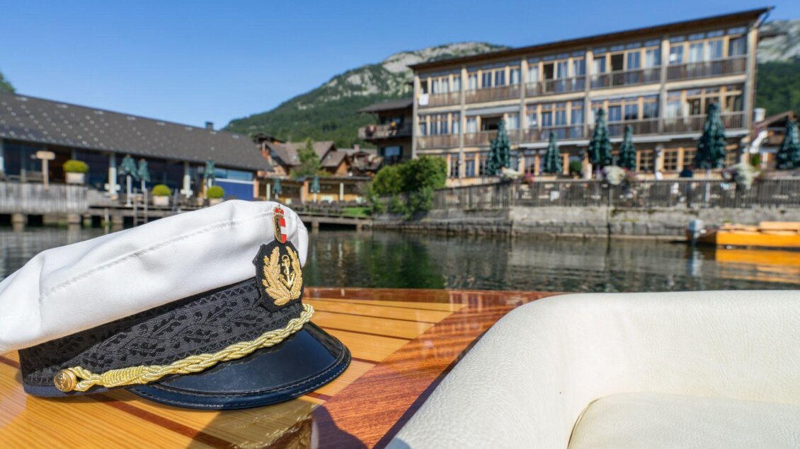 Ein Kapitänshut liegt auf einem Bootstisch mit Blick auf ein malerisches Ufer und ein Hotel. Im Hintergrund sind Berge und ein blauer Himmel zu sehen. | © Schifffahrt Grundlsee/Florian Loitzl