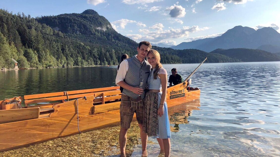 Ein Paar steht am Ufer eines klaren Sees mit Bergen im Hintergrund. Im Wasser ist ein Boot zu sehen. | © Altaussee Schifffahrt