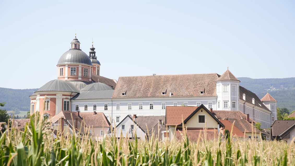 Eine beeindruckende Gebäudeansicht mit einer großen Kuppel und traditionellen Hausdächern im Vordergrund. Im Hintergrund sind Felder mit hohen Pflanzen zu sehen. | © Tourismusverband Oststeiermark