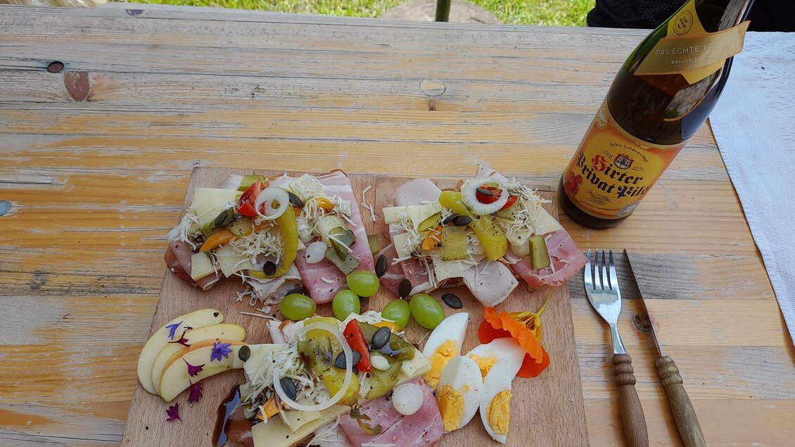 Ein Teller mit verschiedenen Wurst- und Käsesorten, garniert mit Obst und Gemüse. Neben dem Teller steht eine Flasche Bier auf einem Holztisch. | © Brudermann