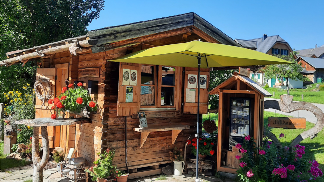 Ein gemütliches Holzhaus mit bunten Blumen und einem Sonnenschirm. Es liegt in einer grünen Umgebung mit einer einladenden Atmosphäre. | © Butterer Hofladen