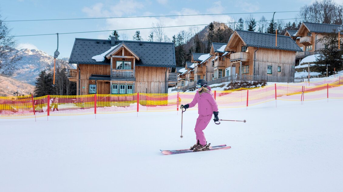 Eine Skifahrerin in pinker Kleidung fährt auf der Piste. Im Hintergrund sind Holzhäuser und verschneite Berge zu sehen. | © www.zloam.at