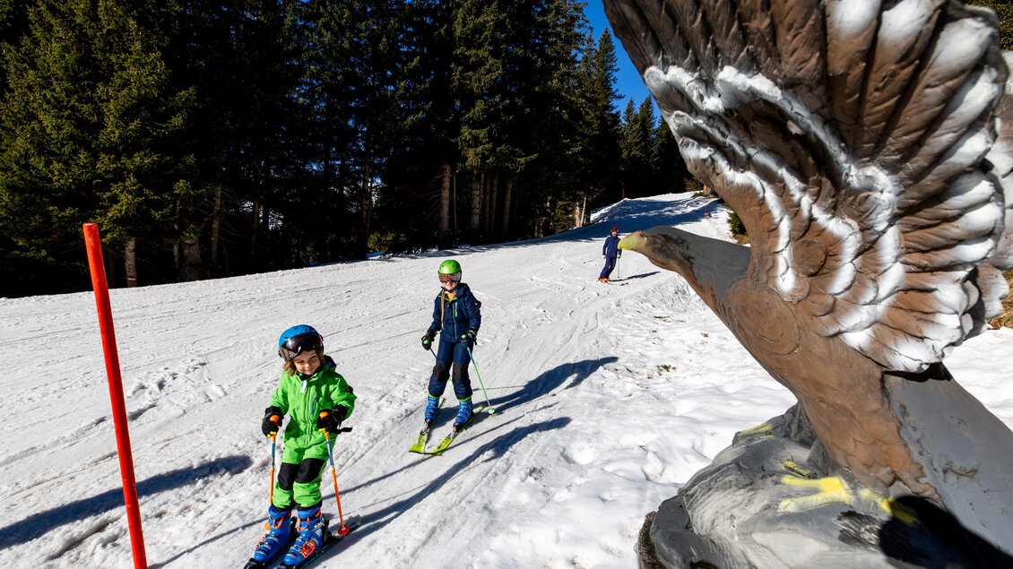 Zwei Kinder fahren Ski auf einer schneebedeckten Piste. Im Hintergrund ist eine große, kunstvolle Skulptur eines Adlers zu sehen. | © Lachtal-Lifte u. Seilbahnen GmbH
