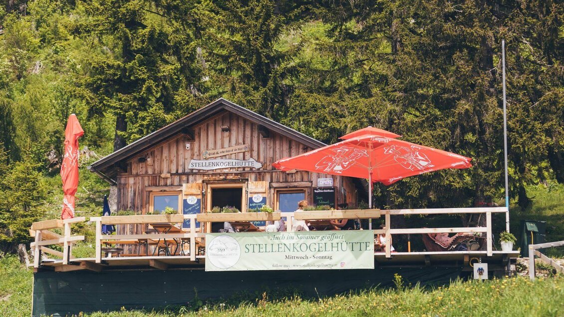 <p>Die Stellenkogelhütte ist eine typische Berghütte mit einer gemütlichen Terrasse und roten Sonnenschirmen. Umgeben von Bäumen und schöner Natur.</p> | © Krewenka