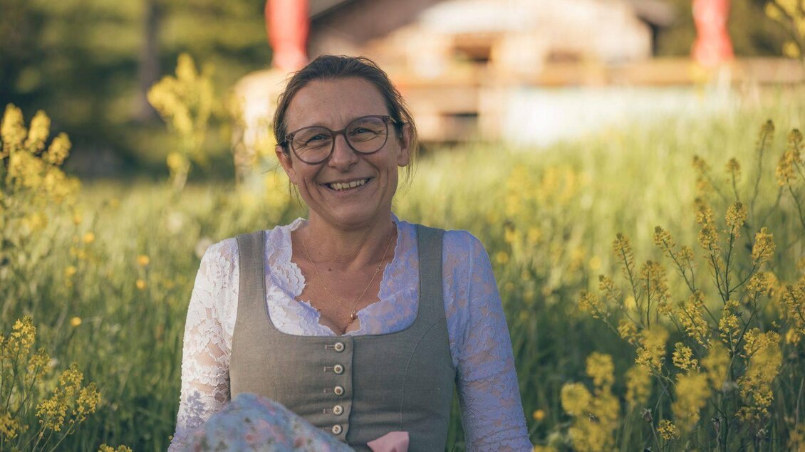 <p><p>Die Wirtin sitzt lächelnd in einen blühenden Feld voller gelber Blumen. Im Hintergrund ist die Stellenkogelhütte  und Bäume zu sehen.</p></p> | © Krewenka