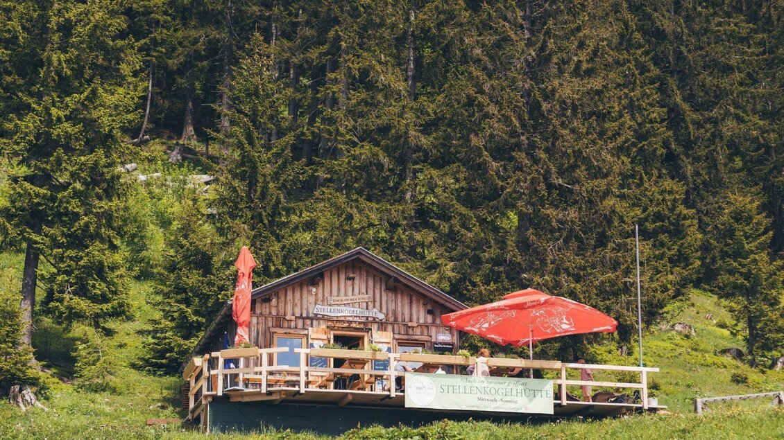 <p>Eine gemütliche Berghütte umgeben von grünen Wäldern. Auf der Terrasse der Stellenkogelhütte stehen Tische mit roten Sonnenschirmen.</p> | © Krewenka
