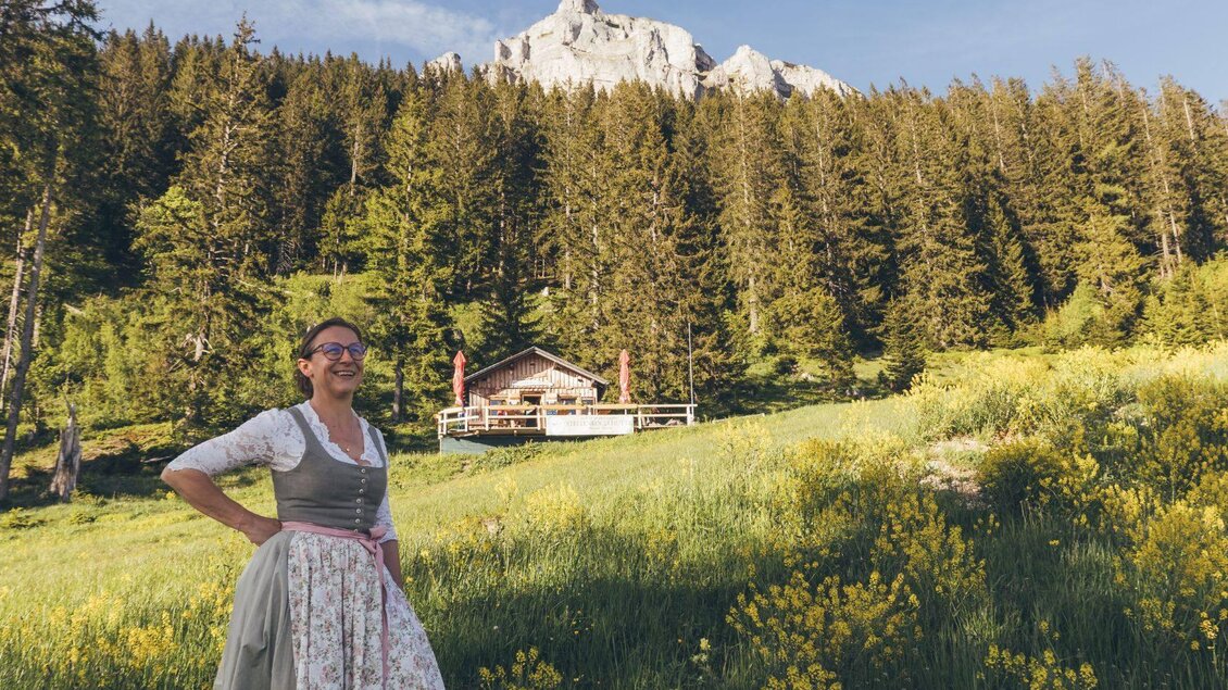 <p>Die lächelnde Wirtin in traditioneller Tracht steht auf einer blühenden Wiese. Im Hintergrund sind Bäume und der Loser zu sehen, sowie die Stellenkogelhütte.</p> | © Krewenka