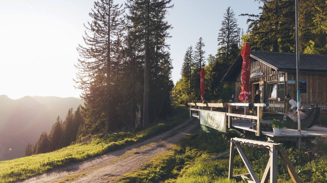<p>Die Stellenkogelhütte am Wanderweg mit Blick auf die Berge. Im Hintergrund stehen hohe Bäume und der Sonnenuntergang sorgt für eine warme Atmosphäre.</p> | © Krewenka