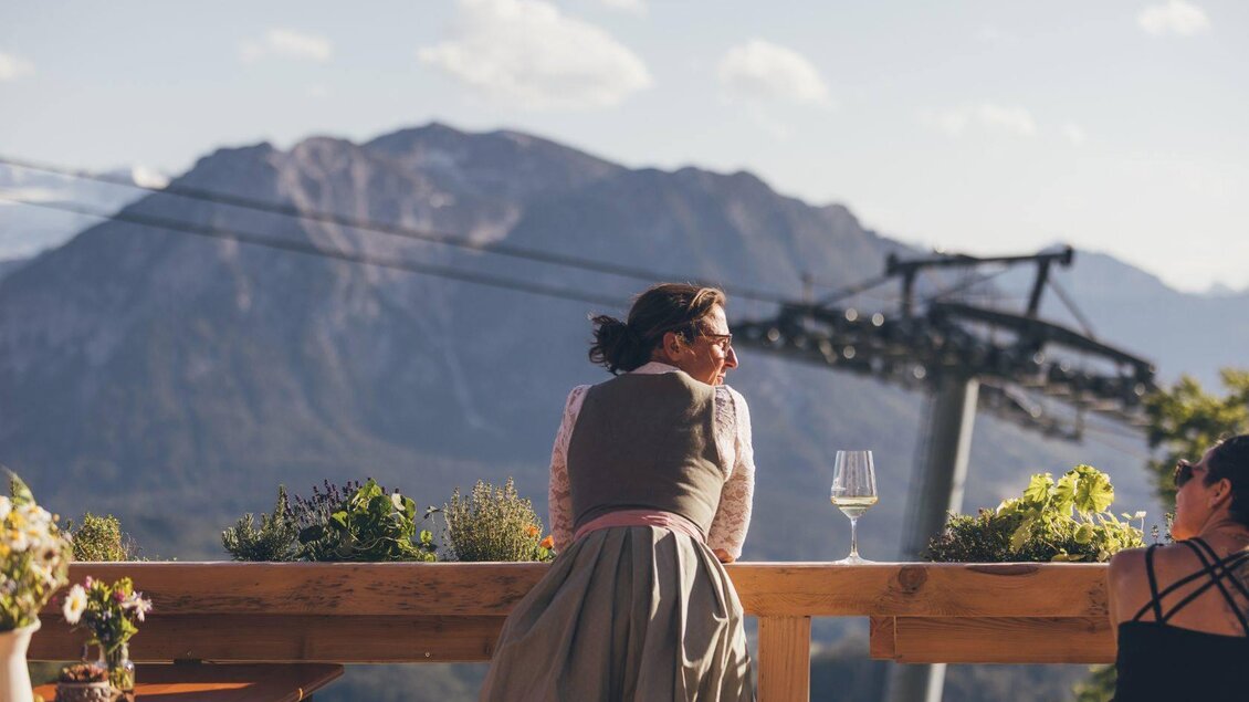 <p>Die Wirtin im traditionellem Dirndl steht am Terassengeländer und blickt Richtung Sandling. Im Hintergrund sind die Loser Panoramabahn, Blumen und der Sarstein zu sehen.</p> | © Krewenka