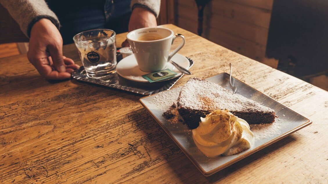 <p><p>Ein Teller mit Schokoladenkuchen und einer Portion Schlagobers werden auf dem Holztisch in der Stellenkogelhütte serviert. Daneben befindet sich eine Tasse Kaffee und ein Glas Wasser.</p></p> | © Krewenka