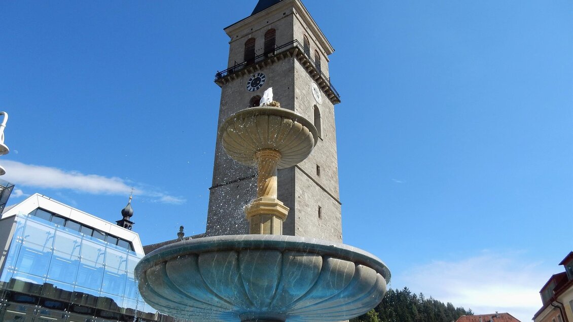 Ein schöner Brunnen im Vordergrund mit einer hohen Uhrturm im Hintergrund. Der Himmel ist klar und blau. | © Planetarium Judenburg
