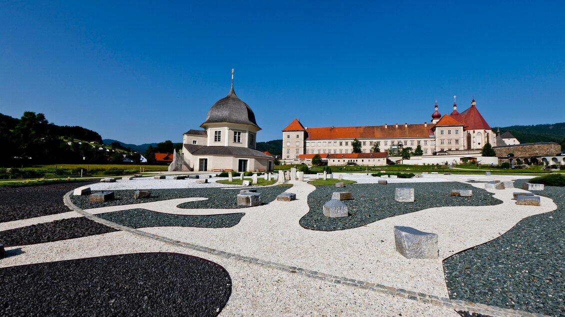 Ein historisches Gebäude mit roten Dächern vor blauem Himmel. Im Vordergrund ist ein schön gestalteter Garten mit Steinen und Kies. | © Holzwelt Murau