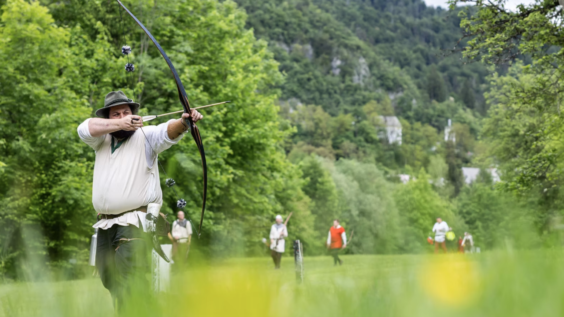 Ein Bogenschütze zielt auf ein Ziel in einer grünen Landschaft. Im Hintergrund sind weitere Personen zu sehen, die ebenfalls am Bogenschießen teilnehmen.