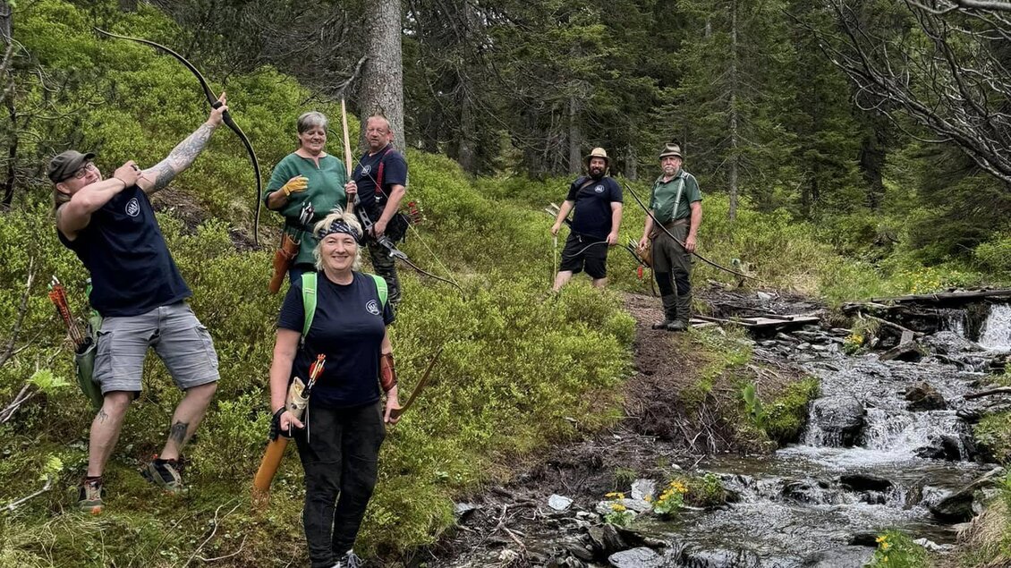 Eine Gruppe von sechs Personen wandert entlang eines schmalen Pfades neben einem kleinen Bach. Die Umgebung ist grün mit vielen Bäumen und einem bewölkten Himmel.
