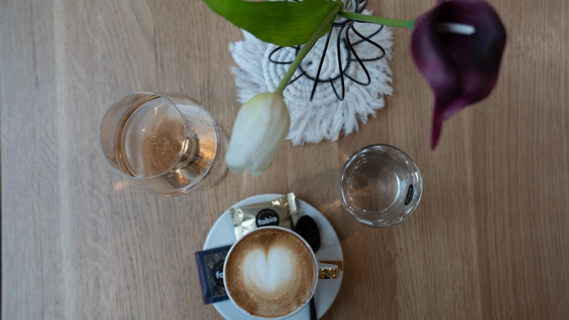 Eine Tasse Cappuccino mit Herz-Muster steht auf einem Teller. Neben der Tasse sind ein Glas Wasser und eine Blume in einer Vase zu sehen. | © STG