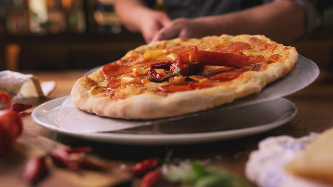 Eine frisch gebackene Pizza mit Paprika und anderen Zutaten wird serviert. Der Hintergrund zeigt eine warme, gesellige Atmosphäre. | © The Kitchen