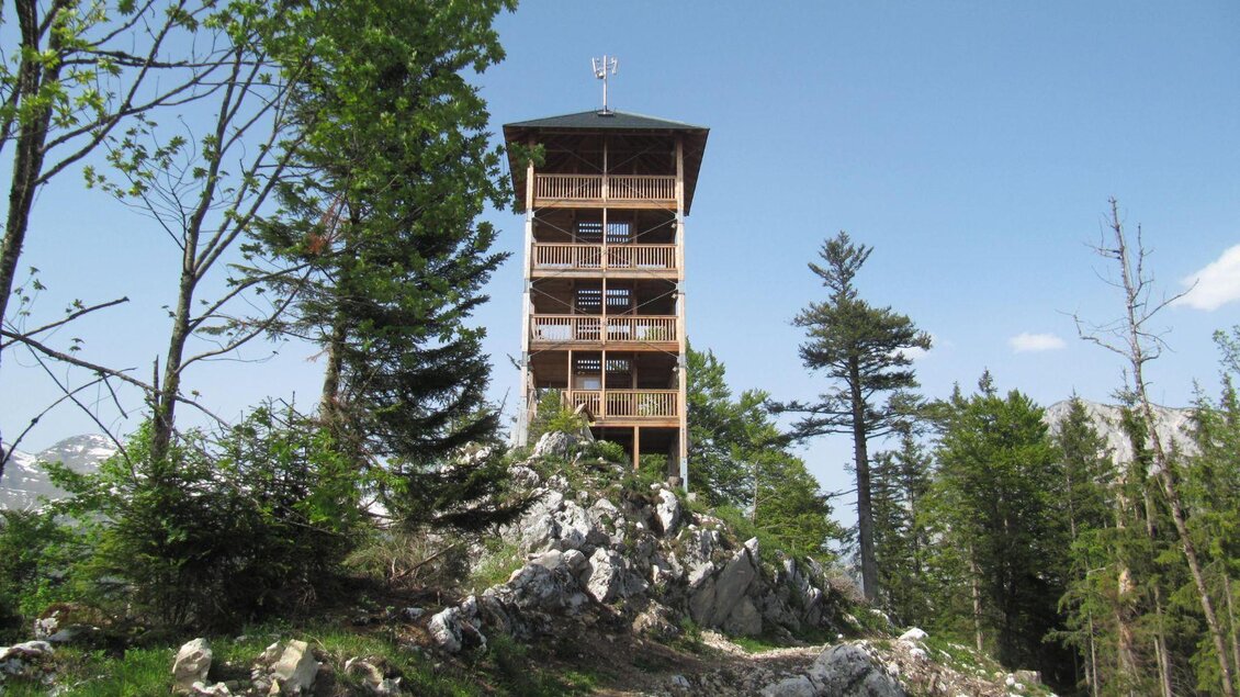 Der Aussichtsturm Tressensteinwarte auf dem gleichnamigen Tressenstein in Bad Aussee bietet eine atemberaubende Aussicht auf die umliegende Bergwelt des Ausseerlandes bis zum Dachstein, sowie auf die umliegenden Seen Grundlsee und Altausseersee. Der Holzturm ragt in den klaren blauen Himmel und ist von tiefgrünen Bäumen und kargen, felsigen Steinen umgeben. | © Wolfgang Kreutzer