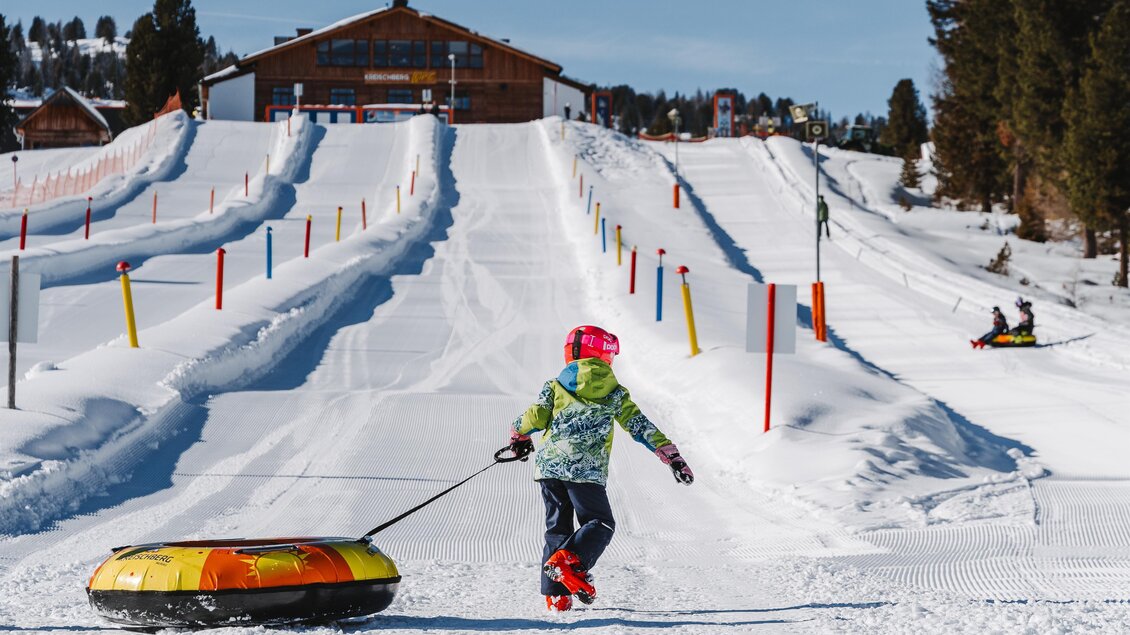 Ein Kind zieht einen Tubes auf einer schneebedeckten Rodelbahn. Im Hintergrund ist ein Holzgebäude und weitere Aktivitäten im Schnee zu sehen. | © TV Murau