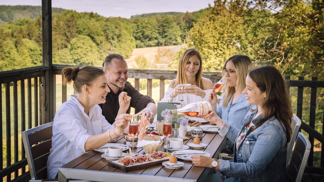 Eine Gruppe von fünf Freunden genießt ein Frühstück im Freien auf einer Terrasse. Im Hintergrund sind Bäume und eine Landschaft sichtbar. | © Vulcano Schinkenmanufaktur
