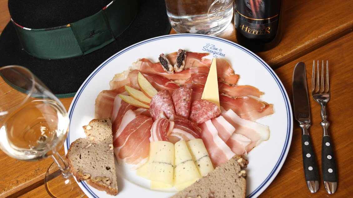 Ein Teller mit einer Auswahl an Wurst, Käse, Obst und Brot. Dazu sind ein Glas Wasser und eine Flasche Wein sowie ein Hut und Besteck zu sehen. | © Vulcano Schinkenmanufaktur