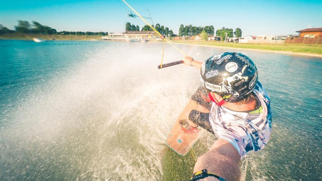 Ein Sportler wakeboardet auf einem klaren Wasser mit spritzenden Wellen. Der Himmel ist blau und sonnig, was die lebendige Atmosphäre unterstreicht. | © Gregor John