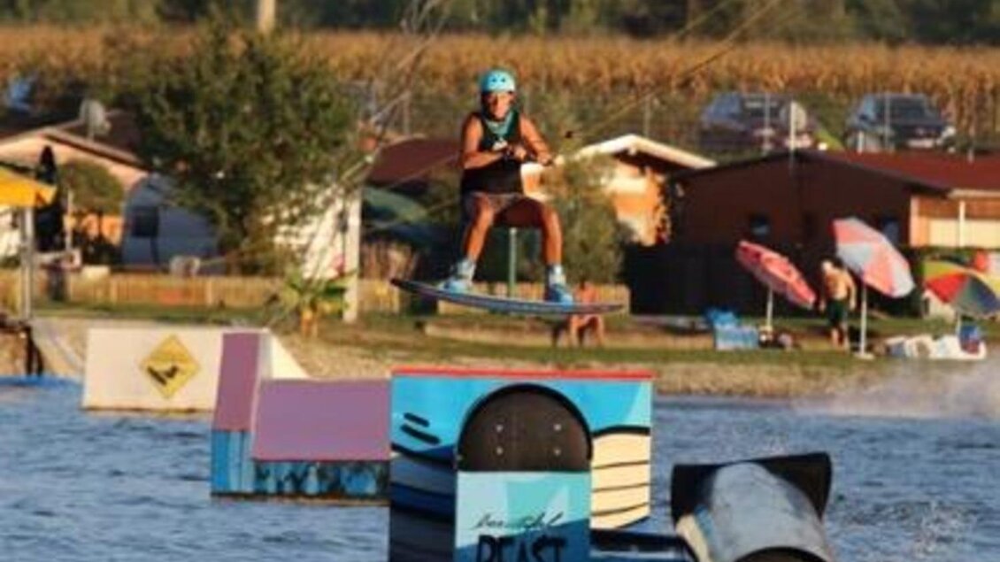 Ein Wakeboarder springt über Hindernisse auf einem Wasserpark. Im Hintergrund sind Bäume und Freizeitboote zu sehen. | © Hermann Paulitsch