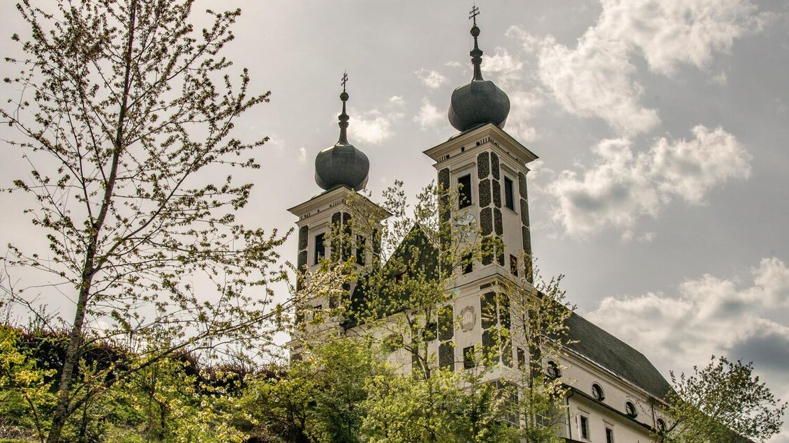 Eine schöne Kirche mit zwei markanten Türmen steht umgeben von Bäumen. Der Himmel ist bewölkt und die Atmosphäre ist ruhig. | © TV Gesäuse