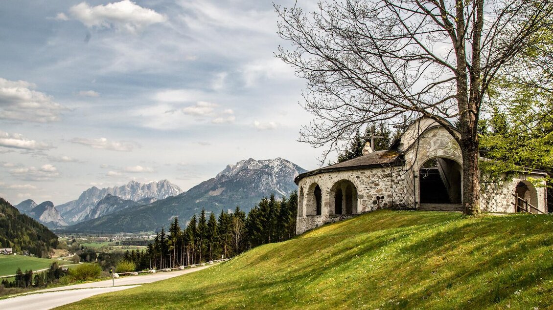 Ein malerisches Landschaftsbild mit Bergen im Hintergrund und einer kleinen, historischen Kirche. Der grüne Hang und der klare Himmel verleihen der Szene eine friedliche Atmosphäre. | © TV Gesäuse