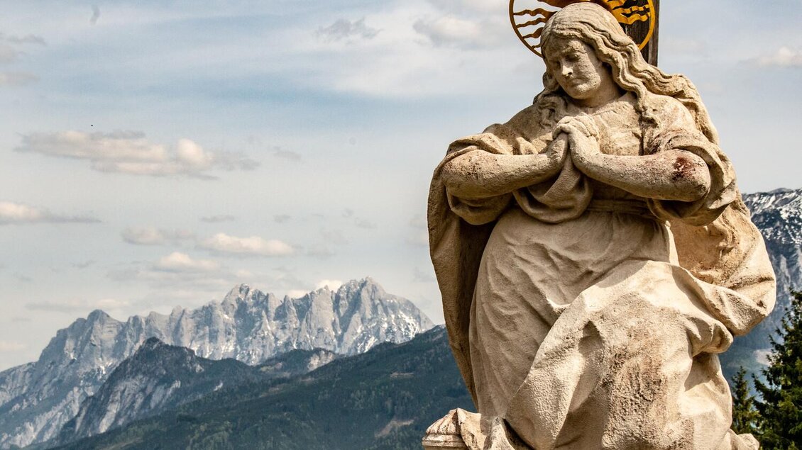 Eine Statue einer Frau mit Heiligenschein steht vor einem beeindruckenden Bergpanorama. Die Szenerie zeigt eine Kombination aus Natur und Kunst. | © TV Gesäuse
