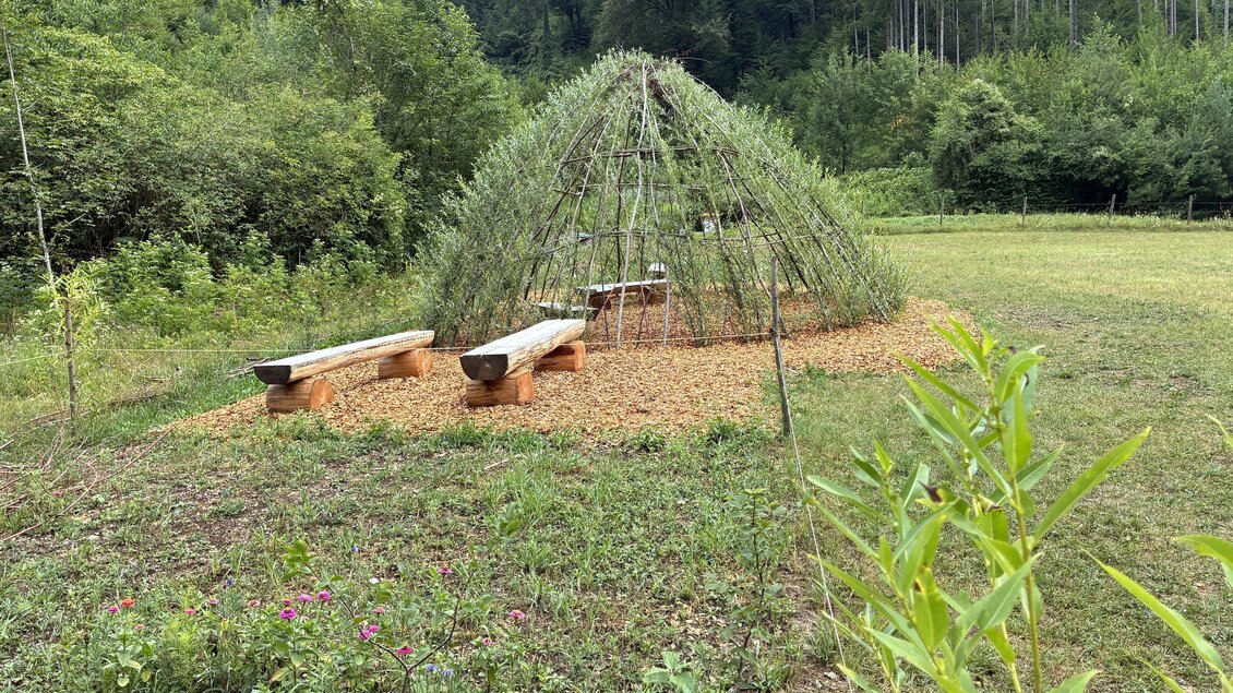 Ein runder, mit Ästen und Pflanzen bedeckter Rückzugsort in einer grünen Umgebung. Daneben stehen zwei Holzbänke auf einem kieseligen Boden. | © Gerd Neuhold
