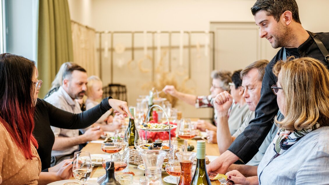 Ein geselliges Essen mit mehreren Personen an einem Tisch. Eine Person serviert Speisen, während andere Gäste essen und anstoßen. | © Der M-Effekt