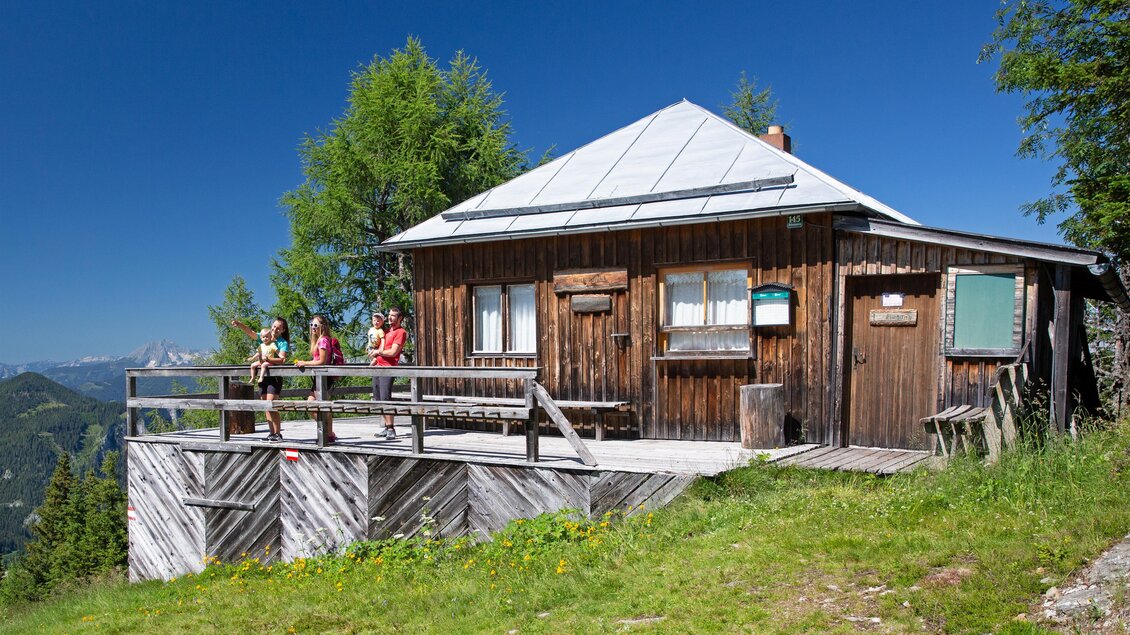 Eine gemütliche Berghütte umgeben von Natur. Drei Personen stehen auf der Veranda und genießen die Aussicht. | © Herbert Raffalt