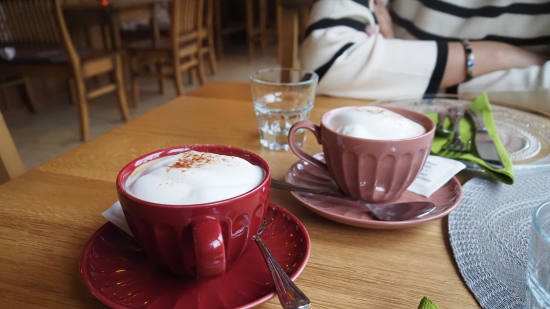 Zwei Tassen mit Cappuccino auf einem Holztisch, eine rot und eine pink. Im Hintergrund sind verschwommen Stühle und ein Glas Wasser zu sehen. | © Tourismusverband Ausseerland