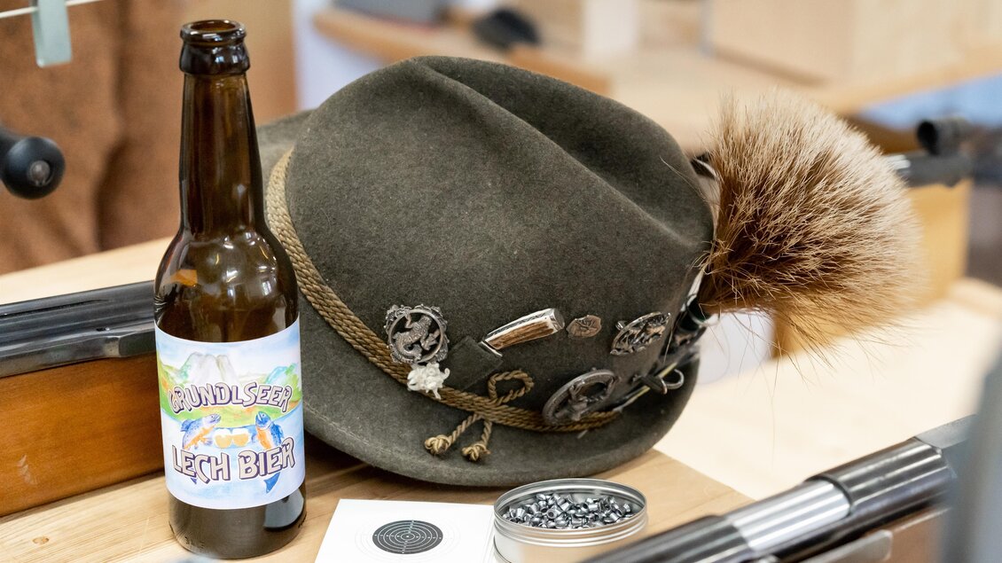 Ein traditioneller Hut mit einer Feder und verschiedenen Ansteckern liegt neben einer Flasche Bier. Im Hintergrund sind Holzmaterialien zu sehen, die eine gemütliche Atmosphäre schaffen. | © www.zloam.at