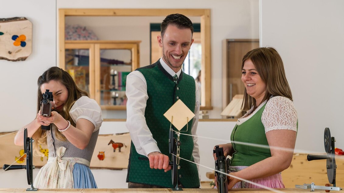 Drei Personen in traditioneller Kleidung stehen an einem Tisch und nehmen an einer Schießaktivität teil. Sie lachen und haben Spaß bei der gemeinsamen Aktivität. | © www.zloam.at