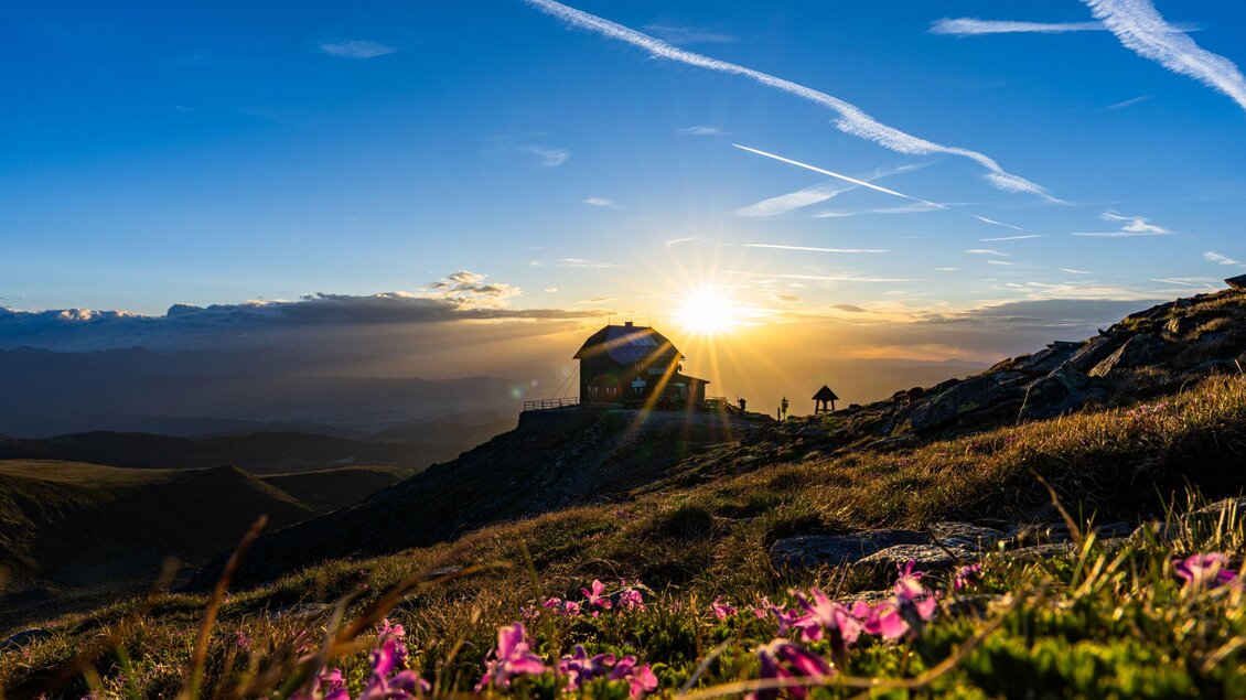 Eine malerische Landschaft mit einem Bauernhaus im Sonnenuntergang. Bunte Blumen schmücken den Vordergrund und der Himmel ist klar und blau. | © Christoph Trois