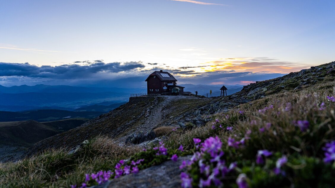 Eine Berghütte steht auf einem Hügel mit einer schönen Aussicht. Im Vordergrund blühen violette Blumen und der Himmel färbt sich bei Sonnenuntergang. | © Christoph Trois