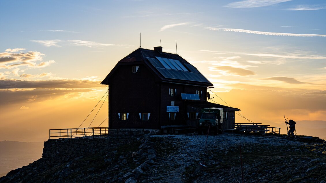Eine Berghütte steht auf einem Gipfel, umgeben von einer dramatischen Abenddämmerung. Der Himmel ist klar mit einigen Wolkenstreifen und es gibt einen Weg, der zur Hütte führt. | © Christoph Trois