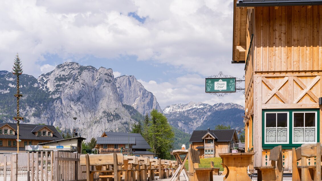Ein charmantes Holzgebäude mit Blick auf beeindruckende Berge. Die Umgebung ist ruhig mit Holzstühlen im Freien und leicht bewölktem Himmel. | © Zloam Wirt, www.zloamwirt.at