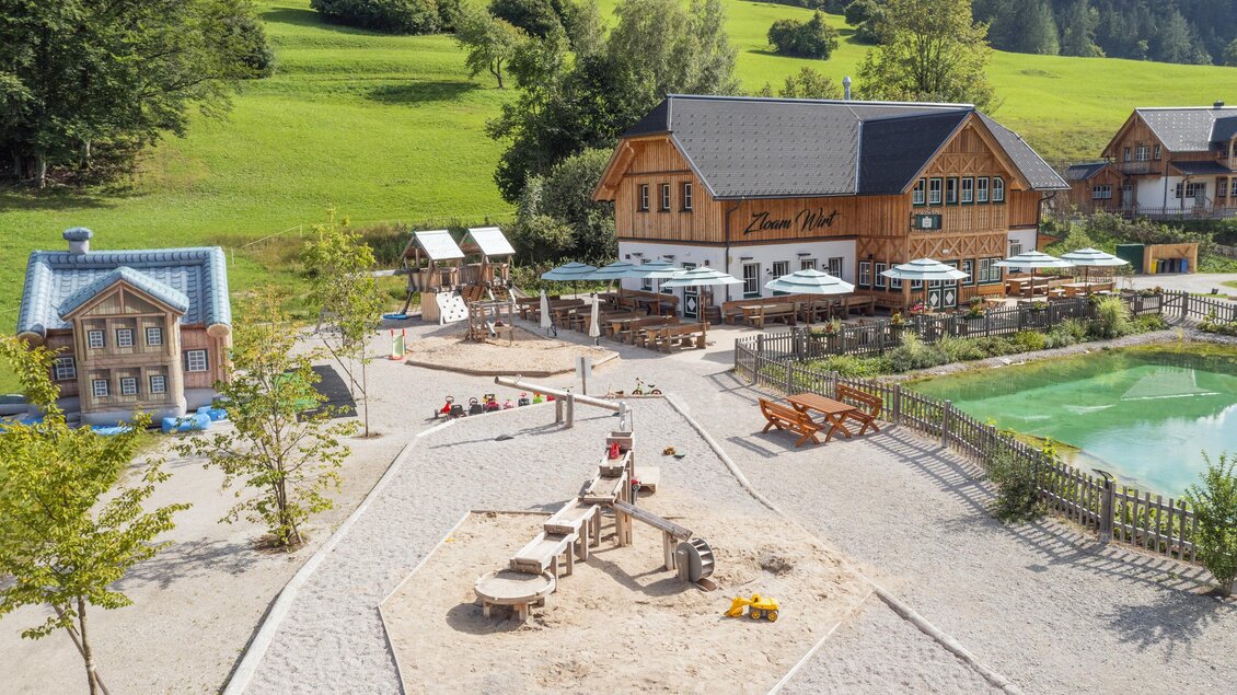 Ein schöner Spielplatz mit einem Sandkasten und einer kleinen Rutsche. Im Hintergrund befindet sich ein gemütliches Restaurant mit Außenplätzen und grüner Landschaft. | © Zloam Wirt, www.zloamwirt.at