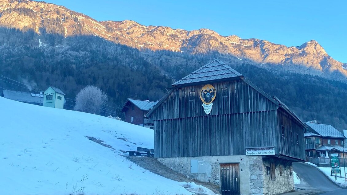 Ein traditionelles Holzhaus mit einem Wappen an der Fassade steht im Vordergrund. Im Hintergrund sind schneebedeckte Berge und ein klarer blauer Himmel zu sehen. | © Bettina Scheck