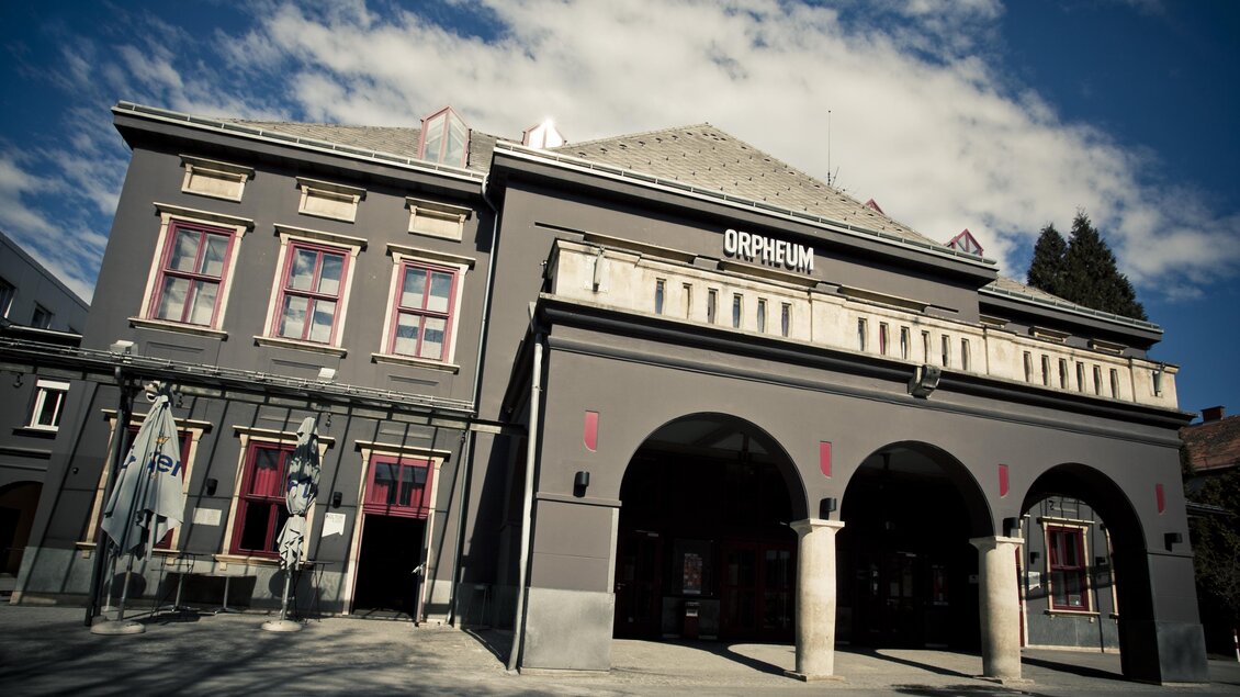 Ein historisches Gebäude mit der Aufschrift "Orpheum". Es hat graue Wände und große Bögen im Eingangsbereich, umgeben von blauem Himmel. | © (c) Spielstätten - Lupi Spuma