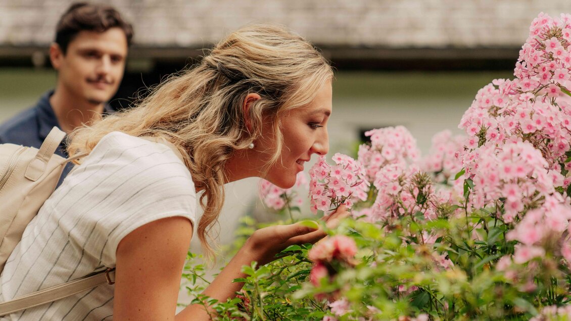 Eine Dame mit blonden Haaren riecht an einer rosa Blume im Bauerngarten im Freilichtmuseum Stübing, im Hintergrund ist ein Mann zu erkennen und ein altes Gebäude. | © Region Graz-studio draussen